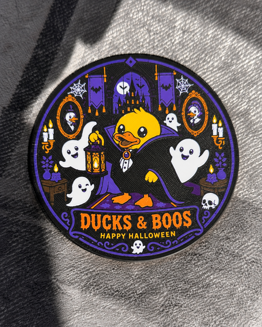A circular coaster featuring a cartoon yellow duck in a black cape with 'DUCKS & BOOS HAPPY HALLOWEEN' text, surrounded by ghosts, bats, candles, and a haunted house on a gray textured fabric background.